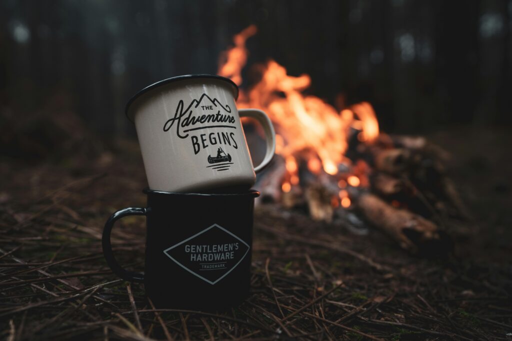 Two mugs with inspirational quotes stacked near a campfire in a tranquil forest environment.