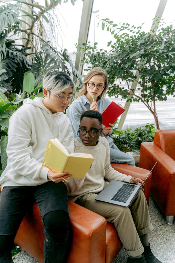 Three diverse students studying and collaborating with books and a laptop indoors.